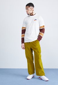 Mladý muž s vitiligom, ktorý má na sebe biele tričko Nike cez dlhé hnedé prúžkované rukávy, žlté cargo nohavice a biele tenisky, stojí na modrej podlahe.