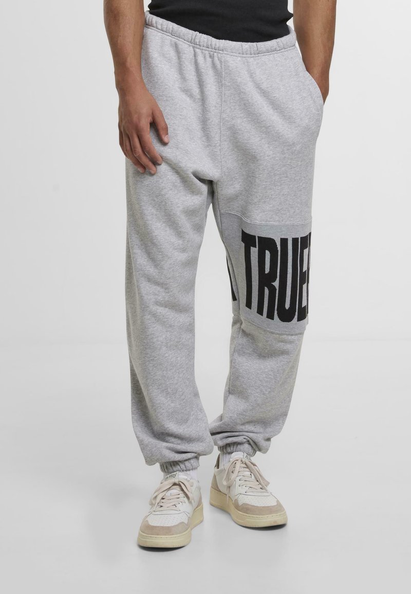 Šedé tepláky s elastickým pásom, sťahovanými členkami a veľkým čiernym nápisom "TRUE" na ľavom stehne. Vyrobené z mäkkej bavlny.