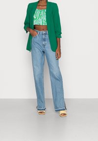 Green blazer over a green and white patterned crop top, paired with light blue wide-leg jeans and yellow block-heeled sandals.