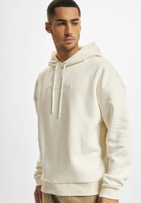Cream hoodie featuring a drawstring, ribbed cuffs and hem, and embroidered text on the front, made from a soft, textured fabric.