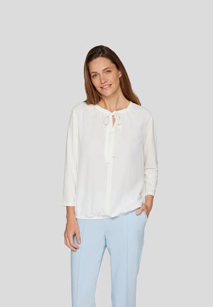 White blouse with 3/4 sleeves and a tie neckline. Soft fabric, slightly gathered waist. Worn with light blue trousers for contrast.