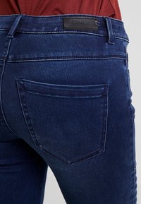 Jeans de mezclilla azul oscuro, con cintura alta, bolsillos traseros y un parche de cuero de la marca en la cintura. Textura suave.