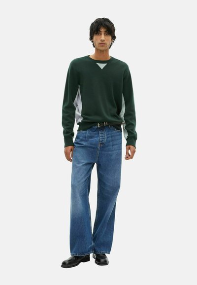Giovane uomo in piedi che indossa un maglione verde scuro con pannelli bianchi ai lati, jeans blu larghi, scarpe nere e una cintura nera, davanti a uno sfondo bianco.