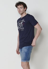 Tumši zils kokvilnas T-krekls ar grafisku apdruku un apaļu kakla izgriezumu, savienots ar gaiši ziliem džinsu šortiem ar sarullētām malām un nolietotām detaļām.