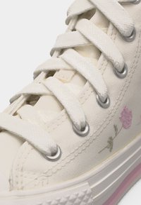 Detailný záber na béžovú plátenú tenisku s bielymi šnúrkami, kovovými očkami, kvetinovou výšivkou a gumovou podrážkou s ružovými pruhmi.