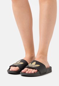 Svarta slidesandaler med en bred rem som har en guld Adidas trefoil-logga. Texturerad sula för grepp, öppet tådesign och minimalistisk stil.