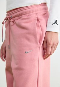 Vaaleanpunaiset puuvillajoggerit, joissa on joustava vyötärö ja naru. Sisältää hopeisen Nike-logon ja sivutaskut. Pehmeä rakenne, rento istuvuus.
