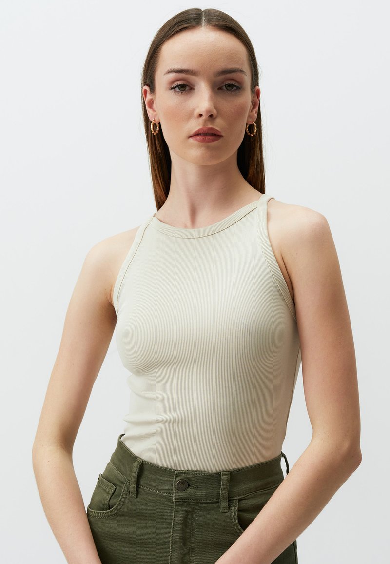 Rippens Struktur beiger Tank-Top mit hohem Kragen und schmalen Trägern, kombiniert mit dunkelgrünen, hochgeschnittenen Hosen mit Seitentaschen.