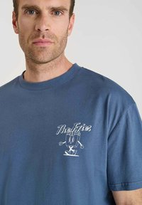 Hombre con una camiseta azul con un personaje de caja de papas fritas de dibujos animados y el texto "Las Papas" en el lado izquierdo del pecho.