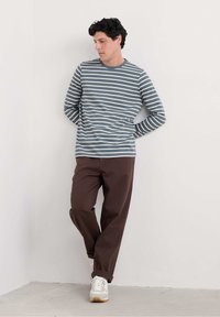 Raidallinen pitkähiha shirt siniharmaana ja valkoisena, yhdistettynä ruskeisiin housuihin. Ominaisuuksiin kuuluu rento istuvuus ja pyöreä pääntie, käytettynä valkoisten tennareiden kanssa.