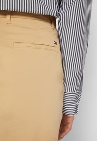 Beige Baumwollhosen mit einer Gesäßtasche und einem kleinen Logo in Rot, Weiß und Blau. Die Bluse hat schwarz-weiße Streifen.