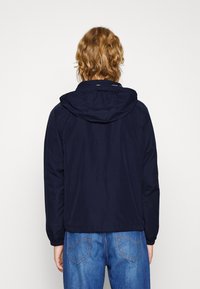 Veste bleu marine avec capuche, fabriquée en matériau léger. Caractéristiques des poignets élastiques et un ourlet légèrement ajusté, avec des détails de couture visibles.
