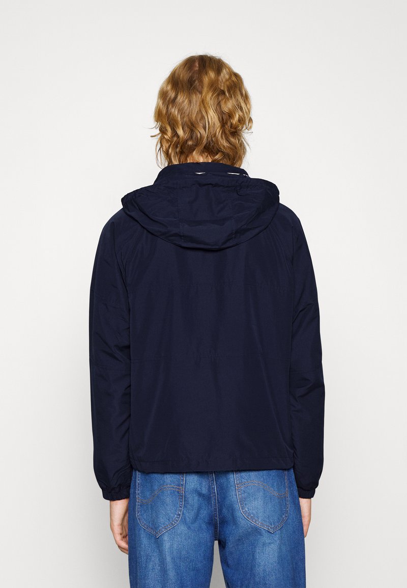Giacca blu navy con cappuccio, realizzata in materiale leggero, presenta polsini elasticizzati e un orlo leggermente svasato, dettagli di cucitura visibili.