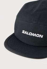 Μαύρο ύφασμα καπέλο με καμπυλωτό γείσο, με λευκή κεντημένη λέξη "SALOMON" στο μπροστινό μέρος.