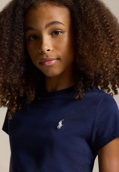 Jaunas žmogus su garbanotais plaukais dėvi tamsiai mėlyną marškinius su baltu išsiuvinėtu polo žaidėjo logotipu ant krūtinės.