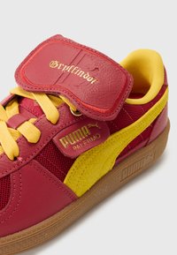 Puma sneakers, piros bőr és hálós felsőrésszel, sárga velúr részletekkel, arany díszítéssel és tépőzáras pánttal, amelyen a "Gryffindor" felirat szerepel.