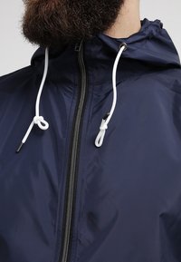 Marinblå vindjacka med dragkedja fram, vita snören och huva. Har ett mjukt, lätt material och svarta dragkedjedetaljer.