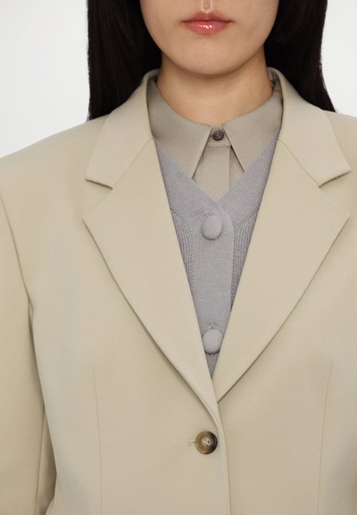 Light beige blazer with a notched lapel, single brown button, layered over a gray knit vest featuring two buttons and texture detail.