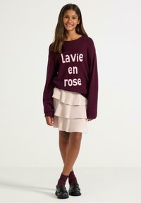 Vínový svetr s bílým nápisem "Lavie en rose", doplněný o vrstvenou béžovou sukni. Černé boty a vínové ponožky celý vzhled dokončují.