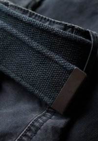 Sort stofbælte med struktureret vævet design, prydet med en metallisk rektangulær spænde, placeret på mørk denim materiale.