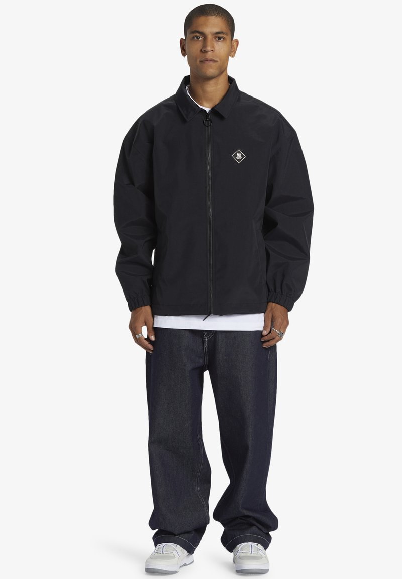 Black, oversized windbreaker jacket with a high collar, zipper front, and elastic cuffs. Paired with dark denim trousers and white sneakers.