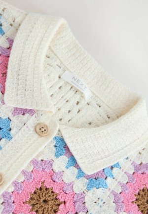 Prim-plan al unei haine croșetate cu guler crem și nasture, având un model floral multicolor în nuanțe de roz, mov, albastru și maro.