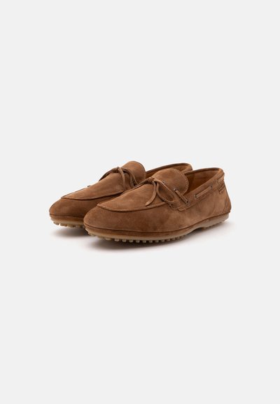 Paire de mocassins en daim marron avec semelles en caoutchouc à picots et détails en lacets sur fond blanc uni.