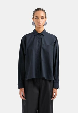 Camicia nera oversize con bottoni, maniche lunghe, taschino sul petto e una superficie liscia. Presenta un colletto classico e tacchi laterali sottili.