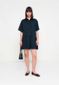 Grünes kariertes kurzärmliges Kleid mit einer Knopfleiste, taillierter Taille und lockerer Passform. Kombiniert mit burgunderfarbenen Ballerinas und einer schwarzen Handtasche.