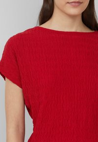 Haut à manches courtes rouge en tissu texturé et ondulé. Encolure ronde avec une coupe décontractée, mettant en avant un motif subtil tout au long.
