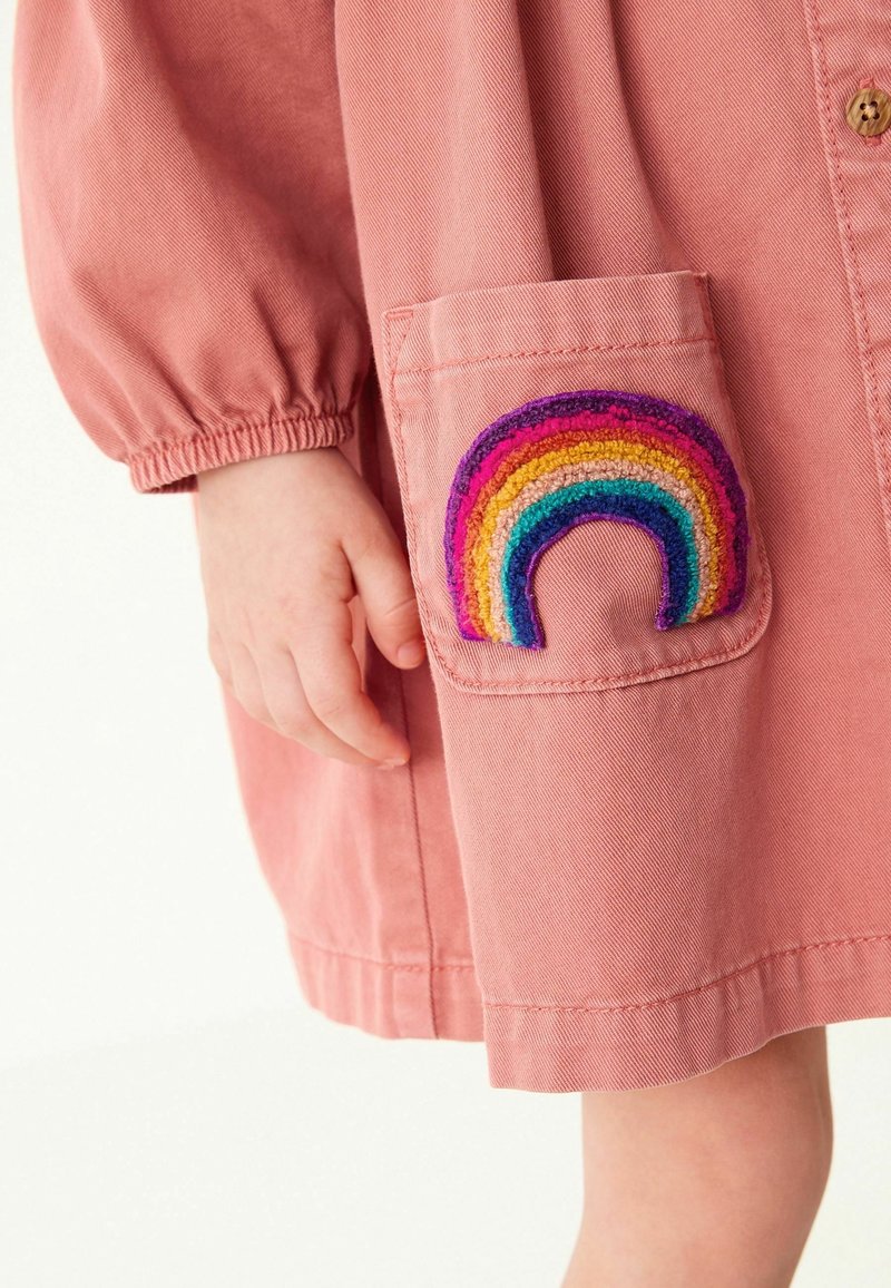 Robe en coton rose avec manches bouffantes et une poche ornée d'un motif arc-en-ciel multicolore brodé. Texture lisse.