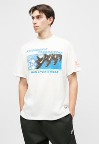 Νέος άνδρας που φοράει λευκό T-shirt της Nike με μπλε γραφικά και το κείμενο «Friendship Tournament» και παπούτσια ποδοσφαίρου, με τα χέρια στις τσέπες μαύρων παντελονιών, κοιτάζει προς τα αριστερά.