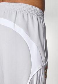 Pantaloni sportivi grigi realizzati in tessuto morbido, con strisce laterali bianche, una cintura elastica e una tasca con zip. Un logo è visibile di lato.