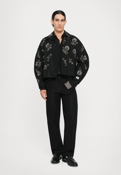 Schwarze, kurz geschnittene Jacke mit Blumenstickerei, kombiniert mit passenden schwarzen Hosen. Beide Teile bestehen aus einem strukturierten Stoff und haben eine schmale Passform.
