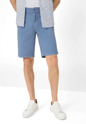 Mann trägt blaue knielange Shorts, weiße Turnschuhe und ein kurzärmeliges, knöpfbares Hemd mit Blumenmuster.