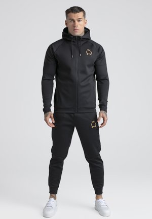 Mann står vendt fremover iført svart hettegenser med glidelås og matchende joggebukser med gullfargede våpenlogoer, kombinert med hvite joggesko.