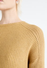 Gros plan d'une personne portant un pull en maille côtelée beige et des boucles d'oreilles anneaux argentés sur un fond clair uni.