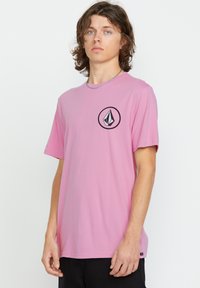 Pink cotton t-shirt with short sleeves, featuring a circular black logo design on the left chest. Crew neckline and relaxed fit.