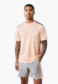 Maglietta sportiva color pesca chiaro con accenti grigi sulle spalle, colletto rotondo e maniche corte. Indossata con pantaloni corti grigi con un pannello colorato laterale.