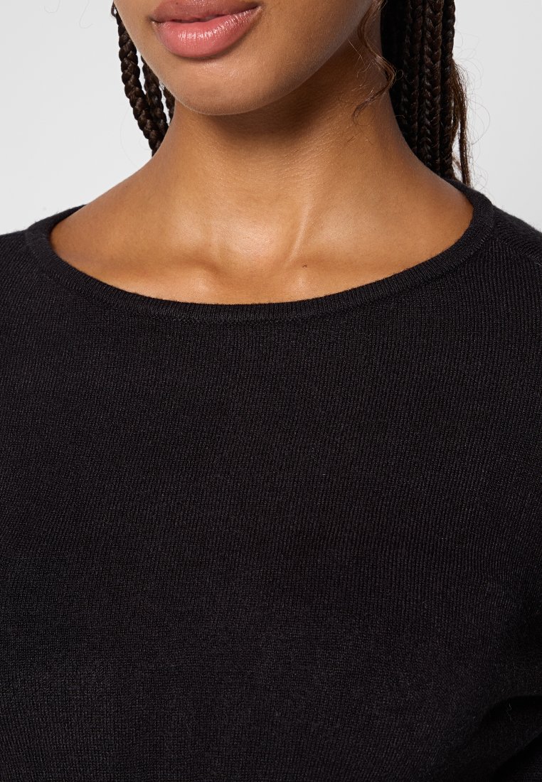 Nahaufnahme einer Person, die einen schwarzen Strickpullover trägt. Ihre geflochtenen Haare und die Lippen in natürlichen Farbtönen sind sichtbar, vor einem neutralen Hintergrund.