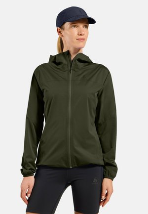 Frau trägt eine dunkelgrüne Jacke mit Reißverschluss und Kapuze, eine marineblaue Kappe und marineblaue Shorts, steht mit entspannt an den Seiten hängenden Händen und schaut nach rechts.