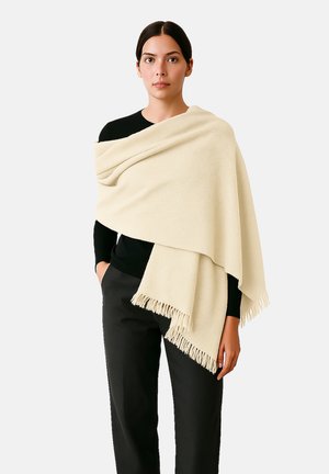 Châle beige en tissu doux, avec des bords frangés, drapé sur les épaules, porté avec un haut noir et un pantalon sombre.