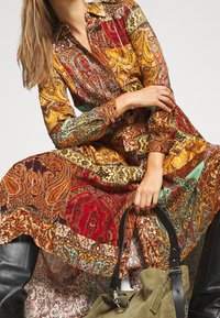 Femme portant une longue robe aux motifs paisley colorés, tenant un sac à main en daim vert olive avec des bottes en cuir noir visibles.