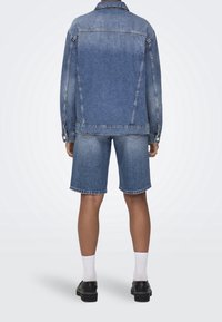 Personne vue de dos portant une veste en jean bleue et un short en jean assorti jusqu'au genou, des chaussettes blanches et des chaussures noires.