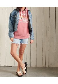 En jeansjacka över en rosa hoodie med grafik, kombinerat med slitna ljusa denimshorts och beige sandaler med en dekorativ rem.
