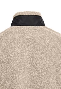 Μπεζ ύφασμα sherpa fleece με υφή κυματιστή επιφάνεια και αντίθετη λείο μαύρο περιλαίμιο στην κορυφή.