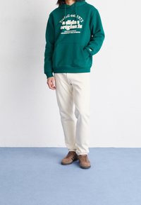 Grön hoodie med vit text och dragsnöre, matchad med beige byxor och bruna skor. Mot en ljus bakgrund med blått golv.