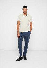 Smėlio spalvos polo marškinėliai su trumpomis rankovėmis ir apykakle. Tamsiai mėlynos siaurėjančios kelnės. Juodi batai. Paprastas dizainas, lygus audinys be matomų raštų.