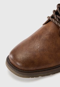 Braune Leder Schnürschuhe mit einer strukturierten Oberfläche, runder Zehenpartie und einer kontrastierenden, helleren Sohle. Ausgestattet mit braunen und beigen Schnürsenkeln.