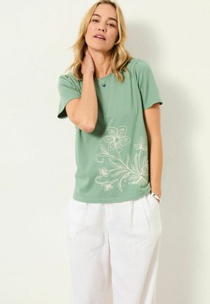 Green cotton t-shirt with short sleeves, featuring white floral embroidery on the lower left. Paired with light-colored, loose trousers.